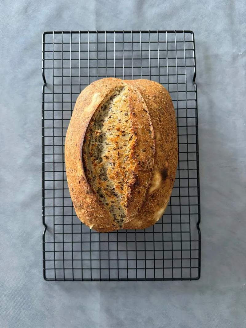 Seeded sourdough loaf with a deep ear, cooling on a rack
